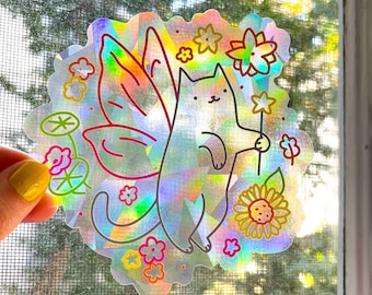 May include: A holographic sticker featuring a cat with butterfly wings, surrounded by flowers and a sunflower. The cat is holding a flower stem in its paw.