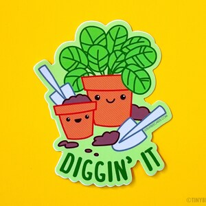 K&ouml;nnte beinhalten: Eine gr&uuml;ne Pflanze in einem Terrakotta-Topf mit einem fr&ouml;hlichen Gesicht, neben einem kleineren Topf mit einem fr&ouml;hlichen Gesicht und einer kleinen Schaufel. Der Text "Diggin' it" steht unter den T&ouml;pfen.