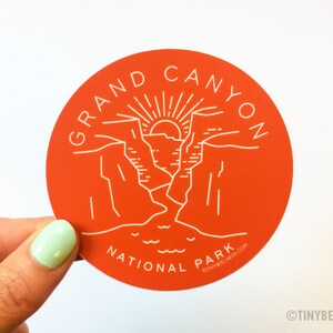 K&ouml;nnte beinhalten: Ein runder, orangefarbener Aufkleber mit einem wei&szlig;en Umriss des Grand Canyon Nationalparks. Der Aufkleber zeigt eine stilisierte Illustration des Canyons mit einer aufgehenden Sonne am Horizont. Der Text "GRAND CANYON NATIONAL PARK" ist um den Rand des Aufklebers geschrieben.