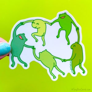 May include: A sticker featuring four green frogs holding hands in a circle. The frogs are different shades of green and have smiling faces. The sticker is on a white background.
