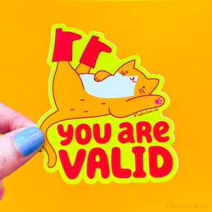 May include: A yellow cat wearing red boots is lying on its back with its paws in the air. The cat is on a bright yellow background with the text "you are valid" in red letters.