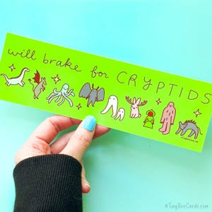 May include: A bright green sticker with the phrase "will brake for CRYPTIDS" and illustrations of various cryptids. The sticker is held by a hand with blue nail polish.