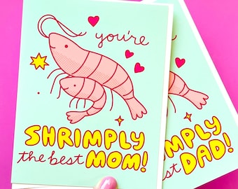 Peut inclure: Une carte bleu clair avec une crevette rose et une plus petite crevette rose sur le dessus. La carte dit "you're shrimply the best mom!" en lettres jaunes avec des accents roses.