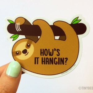 May include: A brown sloth hanging upside down from a branch with green leaves. The sloth has a cute face and the text "How's it hangin?" is written below it.