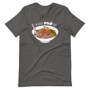Op de afbeelding: Een donkergrijs T-shirt met een witte kom pho erop. De kom bevat noedels, vlees, groenten en een limoenwig. De tekst "I'm crazy PHO you!" is op het shirt gedrukt.