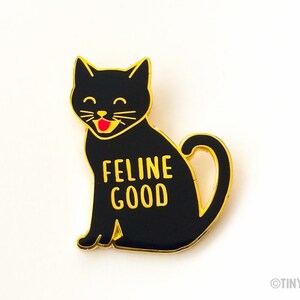 May include: Black enamel pin with a gold outline of a black cat with a red tongue sticking out. The cat is sitting with its tail curled around its body. The text "FELINE GOOD" is written in gold letters on the cat's body.