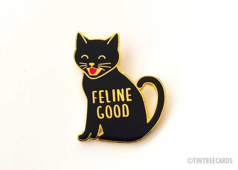 May include: Black enamel pin with a gold outline of a black cat with a red tongue sticking out. The cat is sitting with its tail curled around its body. The text "FELINE GOOD" is written in gold letters on the cat's body.