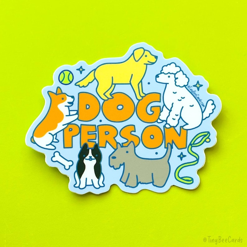 May include: A blue and white sticker with the text "DOG PERSON" in orange letters. The sticker features illustrations of five dogs: a golden retriever, a poodle, a corgi, a black and white dog, and a terrier.