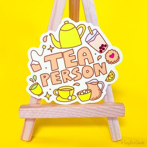 Puede incluir: Pegatina blanca con una tetera amarilla, una taza de t&eacute; y t&eacute; de burbujas. La pegatina dice "TEA PERSON" en letras naranjas.