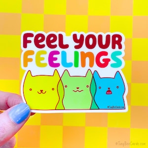 May include: A sticker with three cartoon cats in a row. The cats are yellow, green, and blue. The text "Feel your Feelings" is above the cats in rainbow colors.