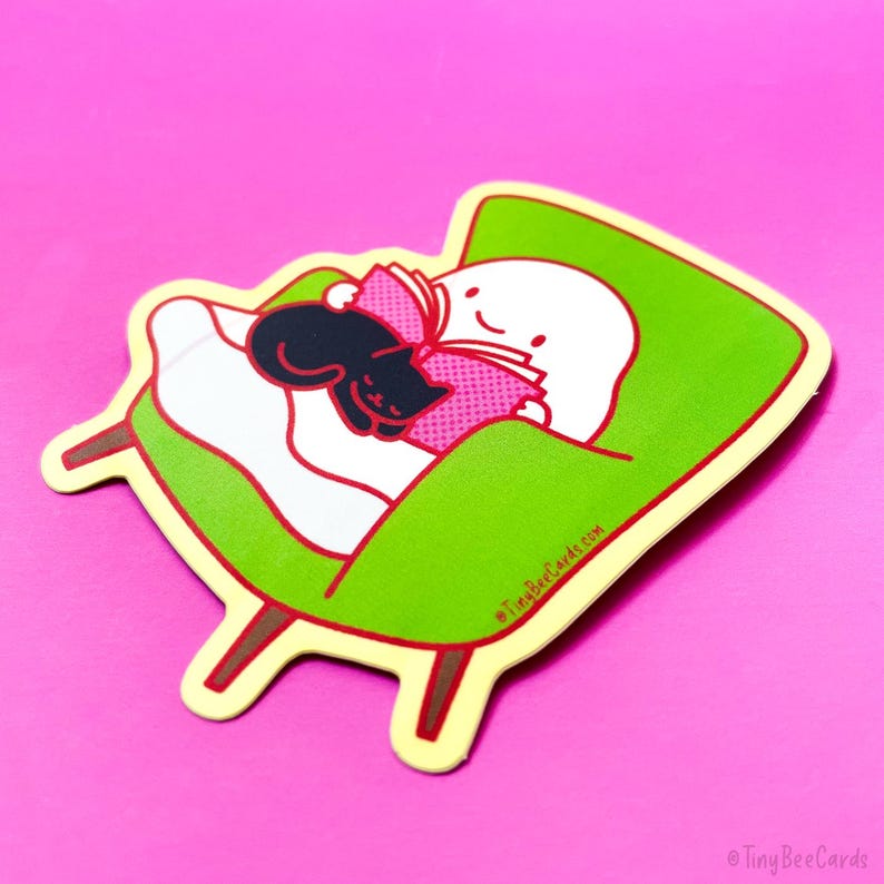 May include: A sticker of a white, round character with a black cat sleeping on its lap. The character is sitting in a green armchair with a pink book. The sticker has a yellow outline and is on a pink background. &copy; TinyBeeCards.com