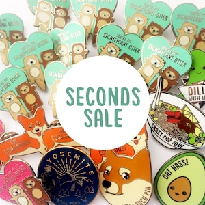 Peut inclure: Une collection de broches en émail avec différents motifs, notamment des animaux, de la nourriture et des scènes de nature. Les broches sont disposées en cercle autour d'un cercle blanc avec le texte "SECONDS SALE" en lettres vertes.
