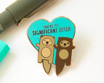 May include: Gold heart-shaped pin with two cartoon otters holding hands. The otters are brown and tan. The text on the pin reads "You're my significant otter."