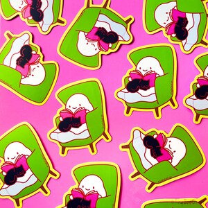 May include: A set of nine stickers featuring a ghost reading a book with a black cat curled up on its lap. The stickers are on a pink background and are in various poses, including sitting in a chair, lying in bed, and reading in a hammock. The stickers are all green and yellow with pink accents.