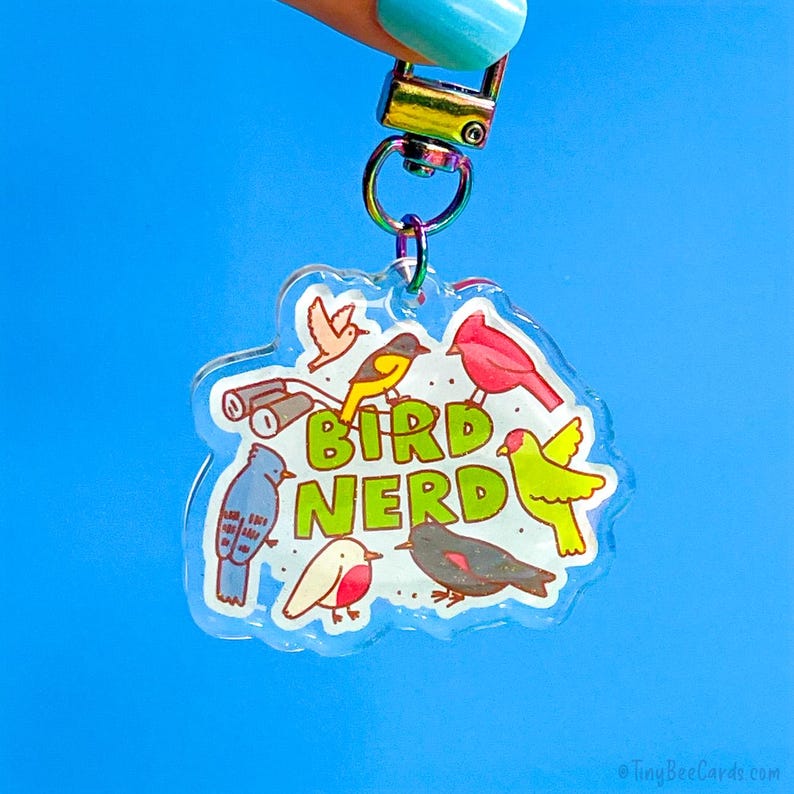 May include: A clear acrylic keychain with a colorful bird illustration and the words "BIRD NERD" in green. The keychain features various bird species and binoculars, attached to a silver-colored clasp.