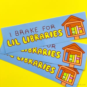 May include: Light blue bumper stickers with the text "I Brake for Lil Libraries" in yellow and red. Each sticker features a cartoon illustration of a miniature library with colorful books. The stickers are on a yellow background.