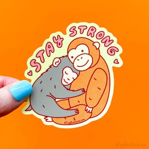 May include: A sticker with an orange background features two cartoon monkeys hugging. The text "STAY STRONG" is in pink, outlined in red. The sticker has a light green border and small pink hearts. The sticker is held by a hand with a blue painted nail.