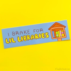 May include: A light blue bumper sticker with the text "I Brake for Lil Libraries" in brown and yellow. A cartoon illustration of a miniature library is on the right side of the sticker.