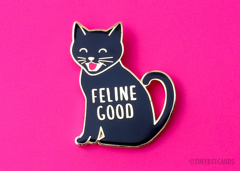 May include: Black enamel pin featuring a cartoon black cat with a white smile and the text "Feline Good" in white letters.