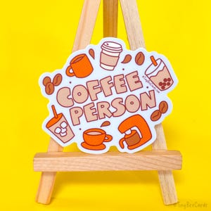 以下が含まれることがあります： コーヒーとオレンジ色の文字で「COFFEE PERSON」と書かれた白いステッカー。ステッカーには、コーヒーカップ、コーヒーポット、ボバティー、コーヒー豆など、コーヒーに関連するさまざまなイラストが描かれています。