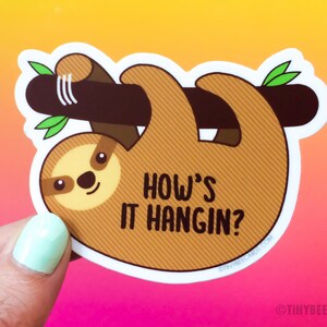 May include: A brown sloth sticker with a white outline, hanging upside down from a branch. The sloth has a cute face and the text "How's it hangin?" is written below it.