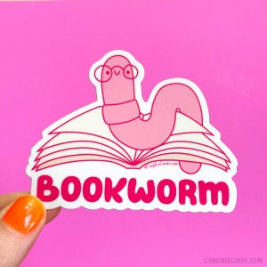 Peut inclure: Autocollant rose en forme de ver de livre avec un ver portant des lunettes et assis sur un livre ouvert. L'autocollant indique "BOOKWORM" en lettres roses.