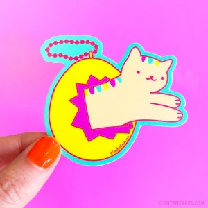 May include: A yellow circle with a pink starburst design and a white cat with yellow and blue stripes peeking out. The cat is wearing a turquoise necklace. The sticker says "@TinyBeecards.com".