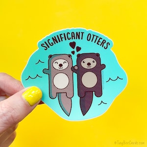 Peut inclure: Un autocollant bleu turquoise avec le texte "Significant Otters" et deux loutres de dessin animé se tenant la main. Les loutres sont grises et violettes avec des ventres blancs et des yeux noirs. Il y a cinq petits cœurs rouges au-dessus des loutres.