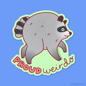 Peut inclure: Un autocollant découpé représentant un raton laveur de dessin animé gris et rond avec une queue rayée, des taches noires autour des yeux et un petit nez rose. Le texte "PROUD weirdo" est écrit en rouge sous le raton laveur.