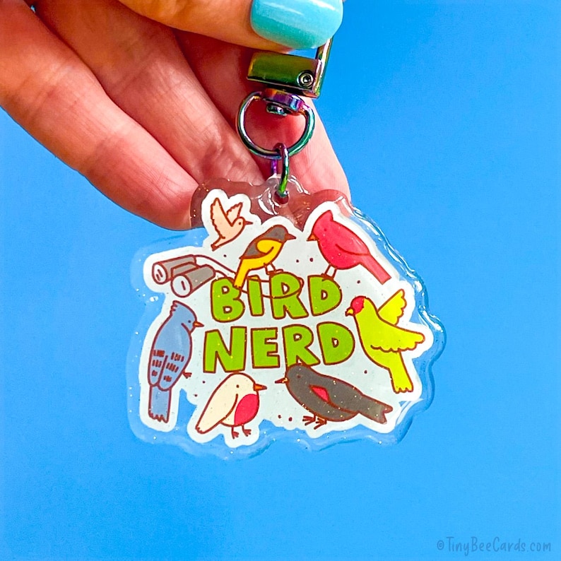May include: A clear, glittery keychain with a colorful bird illustration and the words "Bird Nerd" in green. The keychain features various cartoon birds, binoculars, and a small bird silhouette. The background is a solid blue.