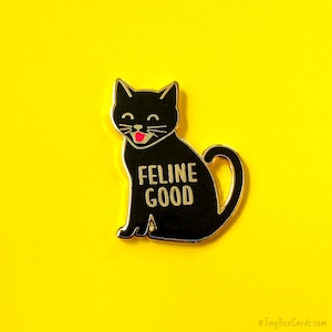 May include: Black enamel pin featuring a cartoon black cat with a pink tongue sticking out. The cat is sitting and has the text "Feline Good" written on its chest.