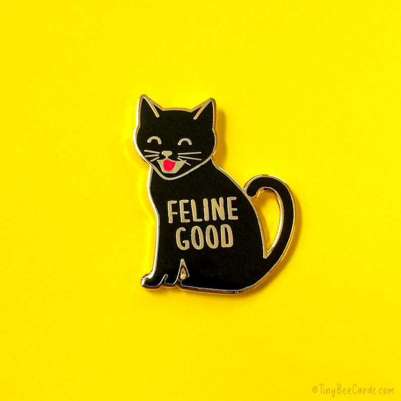 May include: Black enamel pin featuring a cartoon black cat with a pink tongue sticking out. The cat is sitting and has the text "Feline Good" written on its chest.