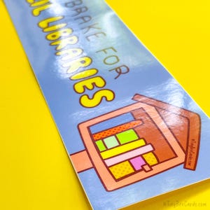 May include: A blue bookmark with the text "BRAKE FOR LIBRARIES" in yellow bubble letters. The bookmark features a brown illustration of a hand holding books with colorful spines. The background is a bright yellow.