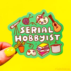 May include: A green sticker with the words "SERIAL HOBBYIST" and illustrations of hobbies like knitting, painting, and playing guitar. The sticker is held against a yellow background.