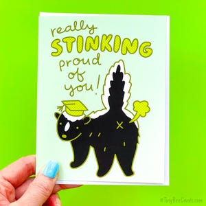 May include: A greeting card with a light green background features a cartoon skunk wearing a graduation cap. The text reads "really STINKING proud of you!" in yellow and green. The skunk's tail is raised, and a yellow cloud emanates from its rear.