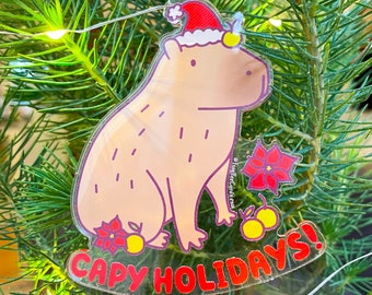May include: A clear plastic Christmas ornament featuring a cartoon capybara wearing a Santa hat. The capybara is sitting down with its legs crossed. The ornament has the text "Capy Holidays!" printed on it in red letters.