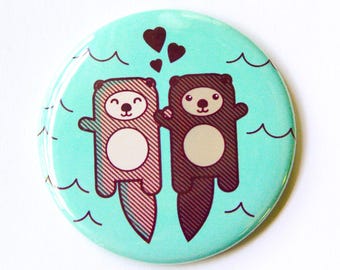 May include: A circular pinback button featuring a light turquoise background with a graphic illustration of two otters holding hands. The otters are brown with diagonal line patterns and white circular bellies. Three dark brown hearts float above.