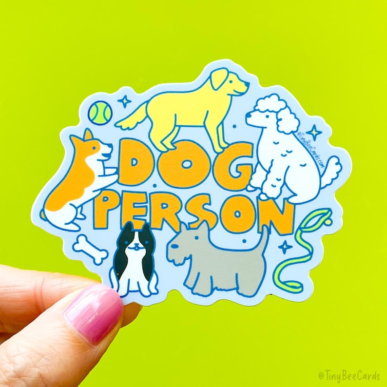 a die cut vinyl sticker that says "dog person" in hand drawn letters, with 5 dogs surrounding the text, including a golden retriever, a white poodle, a corgi, a terrier and a frenchie. They are playing and standing, with toys and bones.