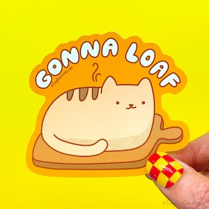 May include: A die-cut sticker with a cartoon cat in a loaf position on a wooden cutting board. The sticker has the words "GONNA LOAF" in an orange bubble. The background is bright yellow.