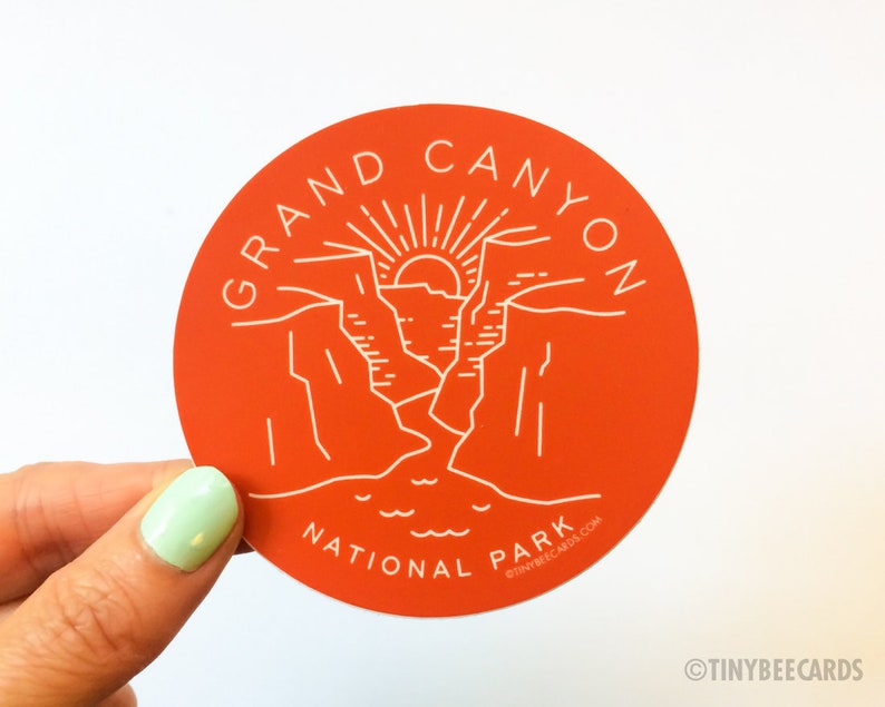 K&ouml;nnte beinhalten: Ein runder, orangefarbener Aufkleber mit einem wei&szlig;en Umriss des Grand Canyon Nationalparks. Der Aufkleber zeigt eine stilisierte Illustration des Canyons mit einer aufgehenden Sonne am Horizont. Der Text "GRAND CANYON NATIONAL PARK" ist um den Rand des Aufklebers geschrieben.