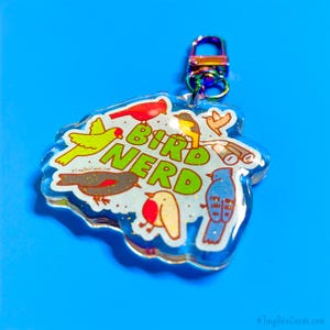 May include: A clear, bird-shaped keychain with the words "Bird Nerd" in green. The keychain features colorful bird illustrations and a rainbow-colored clasp, set against a bright blue background.