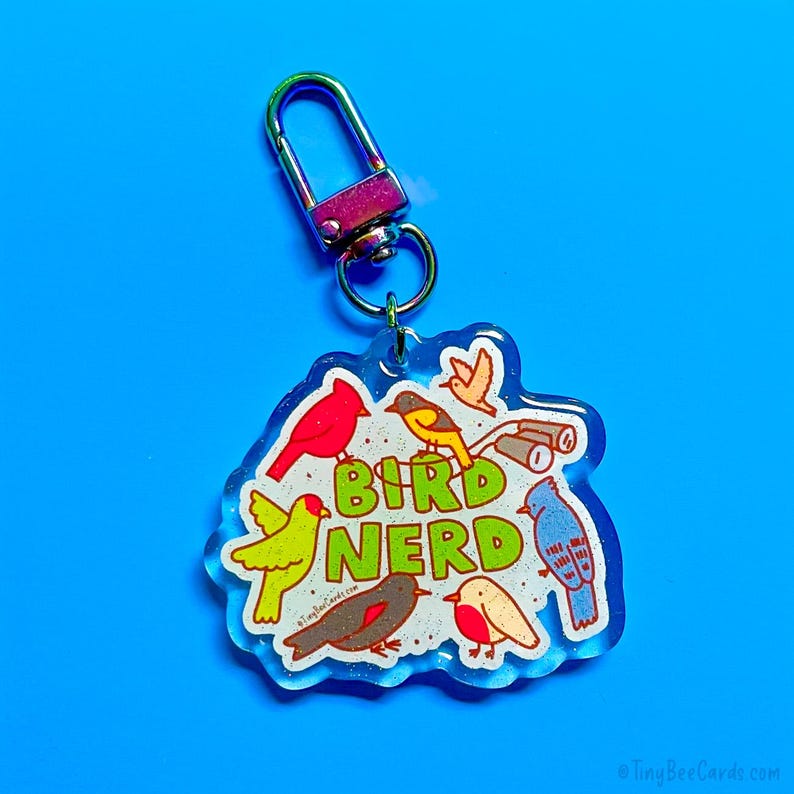 May include: A clear acrylic keychain with a silver clasp. The keychain features a colorful illustration of various birds and the words "Bird Nerd" in green and yellow. The background is a bright blue.