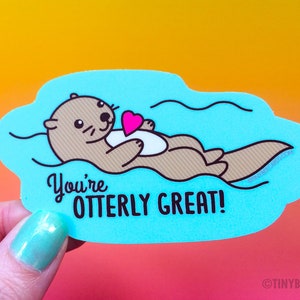 Peut inclure: Un autocollant bleu et blanc avec une loutre de dessin animé flottant sur le dos dans l'eau. La loutre tient un cœur rose. Le texte "You're otter-ly great!" est écrit sous la loutre.