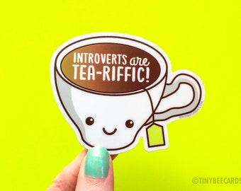 May include: A white teacup with a brown liquid inside and a yellow tea bag hanging from the rim. The teacup has a smiling face and the text "Introverts are tea-riffic!" is written on the cup.