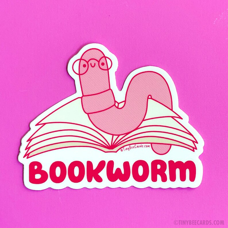 Peut inclure: Un ver de livre rose portant des lunettes est assis sur un livre ouvert avec le texte "BOOKWORM" en lettres rouges.