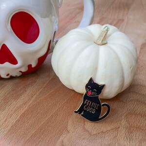 May include: A black enamel pin with a black cat with a red tongue sticking out. The cat is sitting and the text "Feline Good" is below the cat. The pin is on a wooden surface with a white pumpkin and a white ceramic mug with red accents.