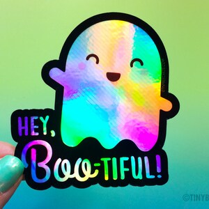 May include: A holographic sticker with a cute ghost design. The ghost is rainbow colored and has a smiling face. The text "Hey, Boo-tiful!" is written in black letters on a black background.