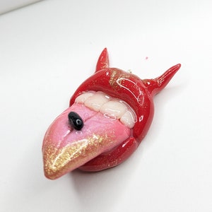 May include: Red and pink clay sculpture of a mouth with a tongue sticking out. The tongue is covered in gold glitter and has a black bead in the center. The mouth has white teeth and red lips. The sculpture is a whimsical and unique piece of art.