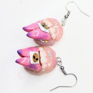 May include: A pair of pink and purple earrings with a glittery finish. Each earring features a sculpted tooth with an alien face sticker on it. The earrings are attached to silver hooks.
