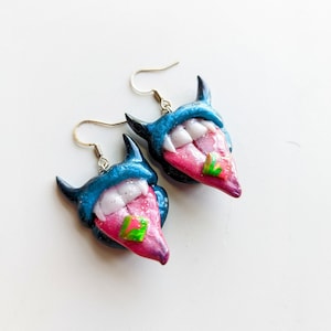 May include: A pair of blue and pink demon earrings with a tongue sticking out and a green and pink design on the tongue. The earrings are made of polymer clay and have silver hooks.